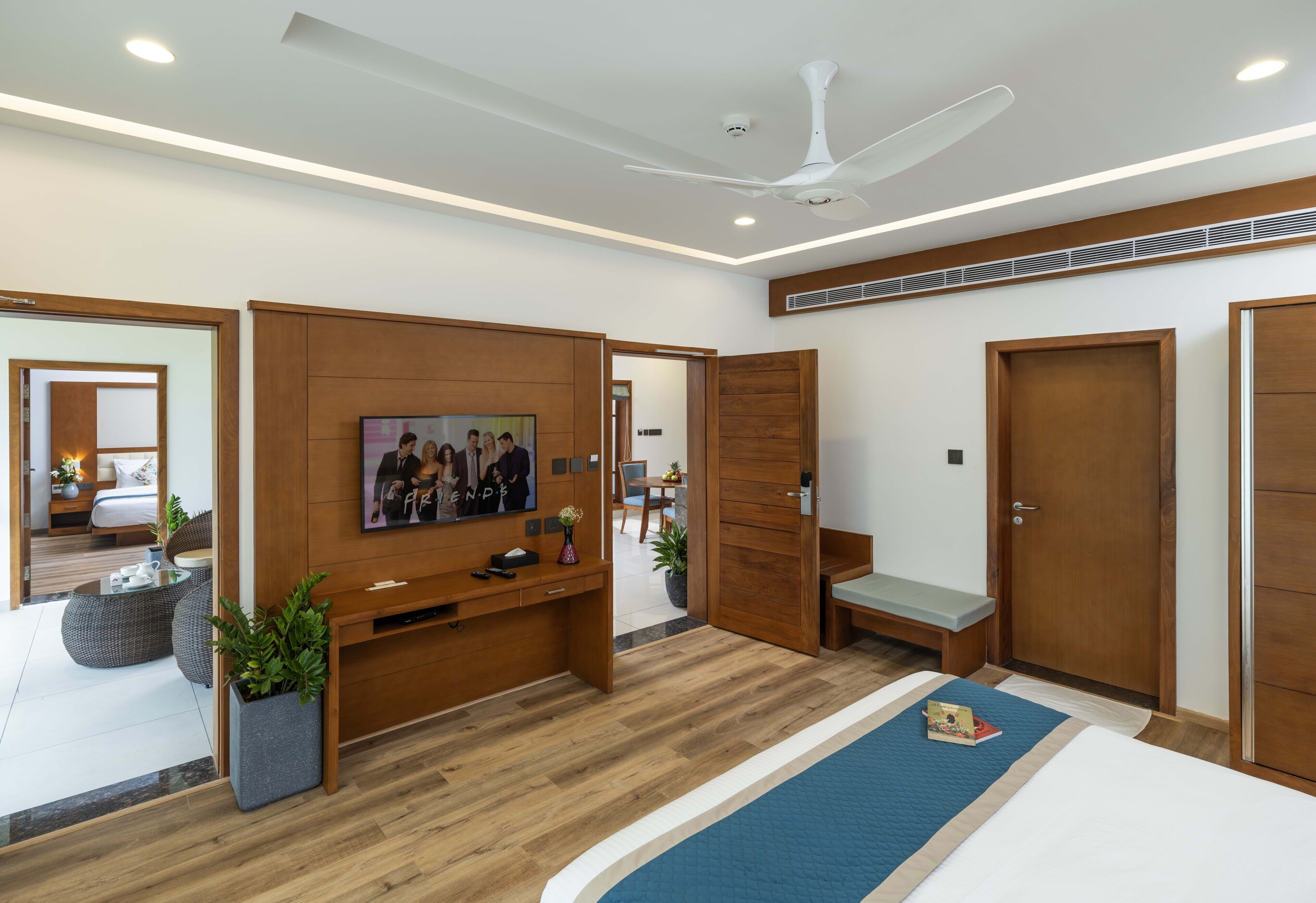 A modern hotel room with a wooden floor, a bed with a blue accent, a TV mounted on the wall, a ceiling fan, and a wooden wardrobe.