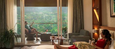 A hotel room with a large bed, a balcony with a view, a sitting area, and a dining table.