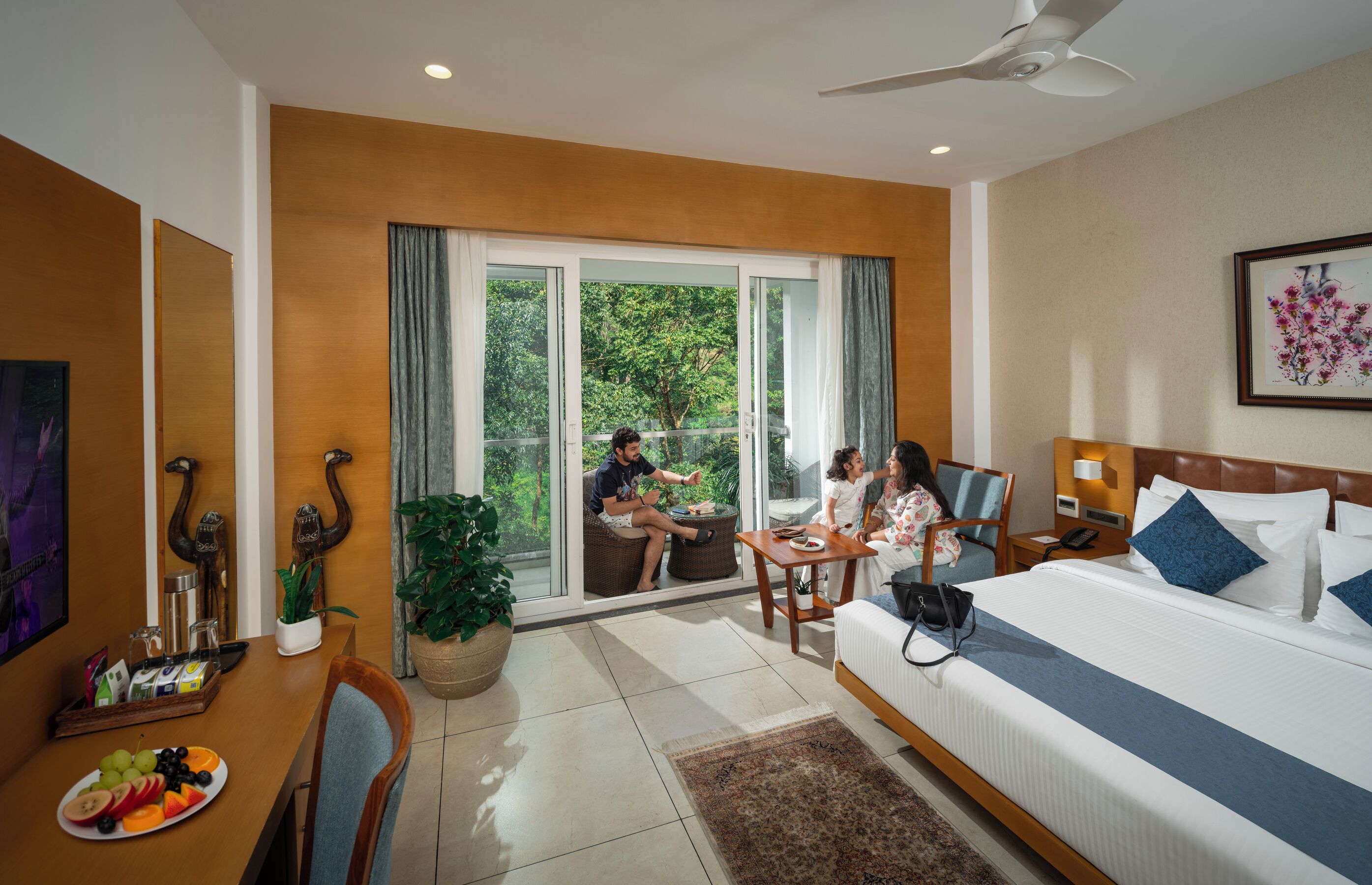A hotel room with a bed, a dining table with fruit, a sofa, a TV, and a balcony with a view of trees.