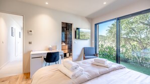 A modern bedroom with a large bed, a desk, and a chair. There is a view of greenery outside the window.