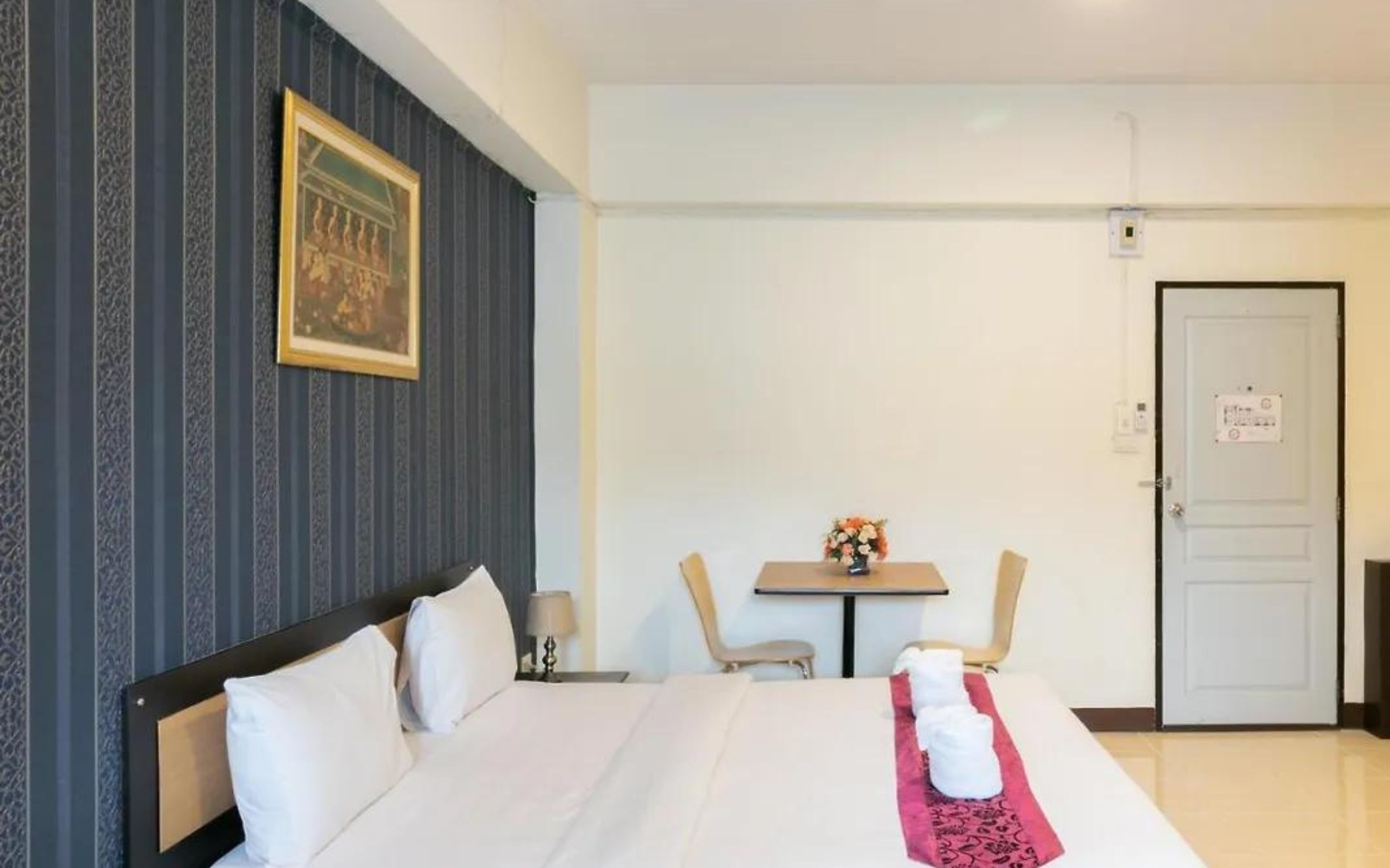 A hotel room with a bed, a small table with a vase of flowers, a chair, and a door with a sign.