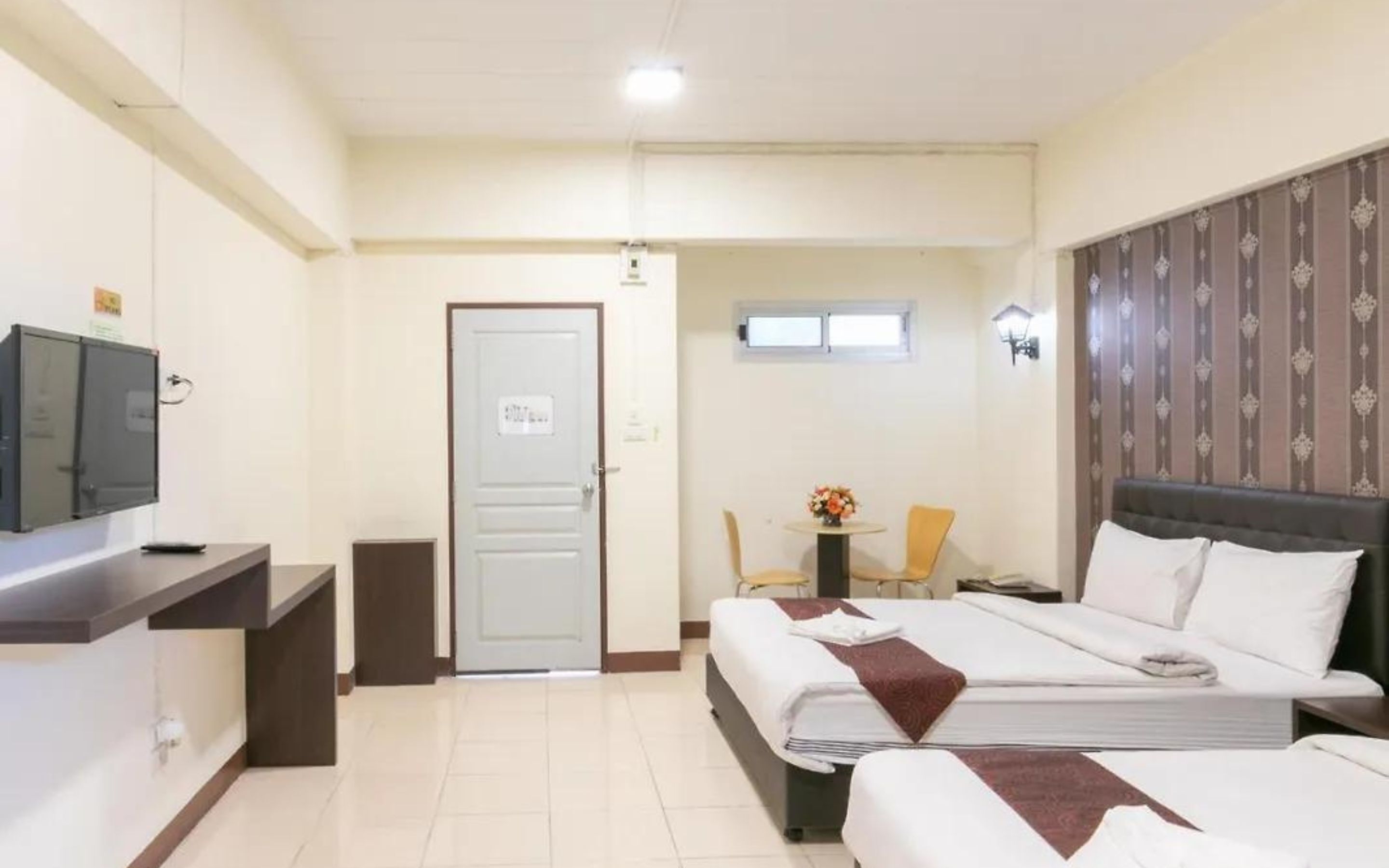 Quarto de hotel com duas camas, uma escrivaninha com computador, uma mesinha com um vaso, e uma porta.