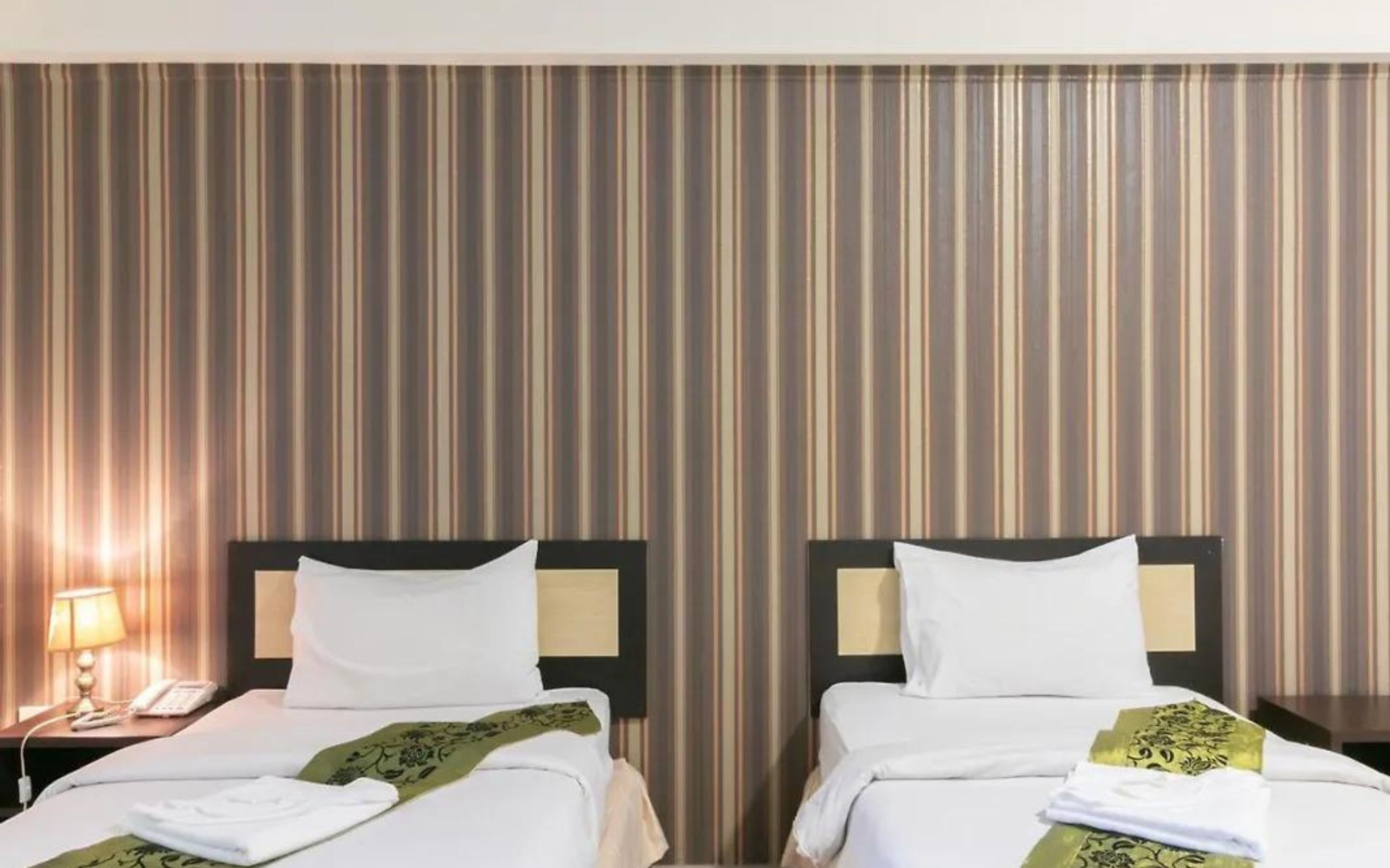 A hotel room with two beds, striped wallpaper, and a nightstand with a lamp and phone.