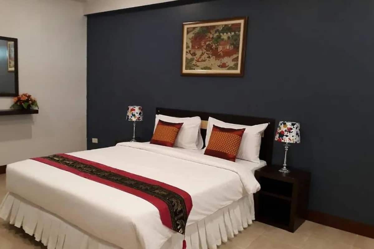 A neatly made bed with white bedding and a red and black decorative border, two bedside lamps with floral patterns, a framed picture on the wall, and a small nightstand with a glass.