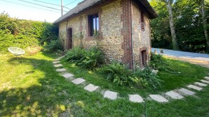 A rustic brick house with a thatched roof, surrounded by lush greenery and a stone pathway leading to the entrance. - La Maison du Garde (STE OPPORTUNE LA MARE)