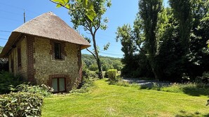A rustic stone building with a thatched roof, surrounded by lush greenery and trees. - La Maison du Garde (STE OPPORTUNE LA MARE)