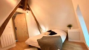A cozy attic bedroom with a sloped ceiling, a wooden door, a bed with white bedding, a fluffy rug, and a small plant on a bedside table. - La Chaumine ` (STE OPPORTUNE LA MARE)