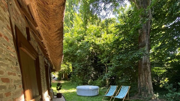 A rustic outdoor area with a stone wall, a thatched roof structure, a small seating area with a cushion, and a tree. - La Chaumine ` (STE OPPORTUNE LA MARE)