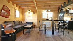 Interior acolhedor de uma cabana, com detalhes em madeira, um sofá, uma mesa de jantar com cadeiras e uma área de cozinha.