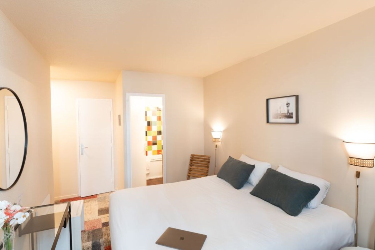 Une chambre d’hôtel avec un grand lit, un bureau avec un ordinateur portable, un miroir et une porte donnant sur une autre pièce.