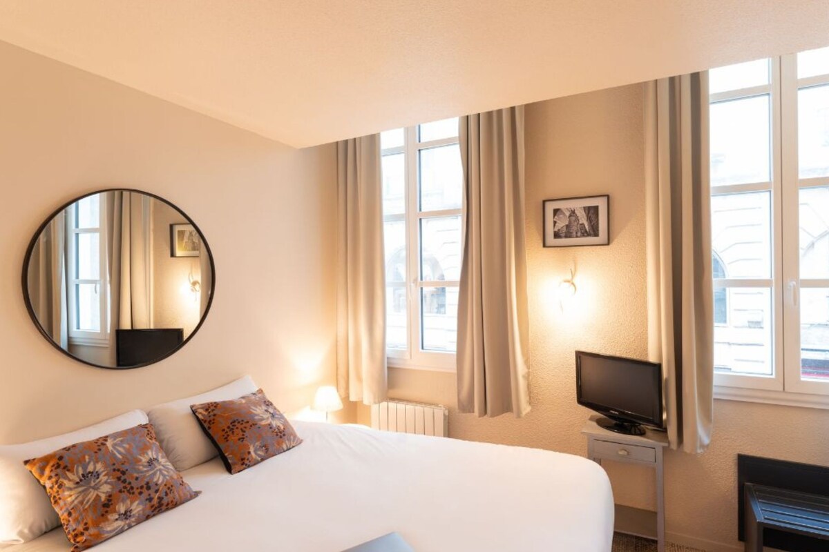 A hotel room with a bed, a television, a desk, a chair, a mirror, and large windows with curtains.