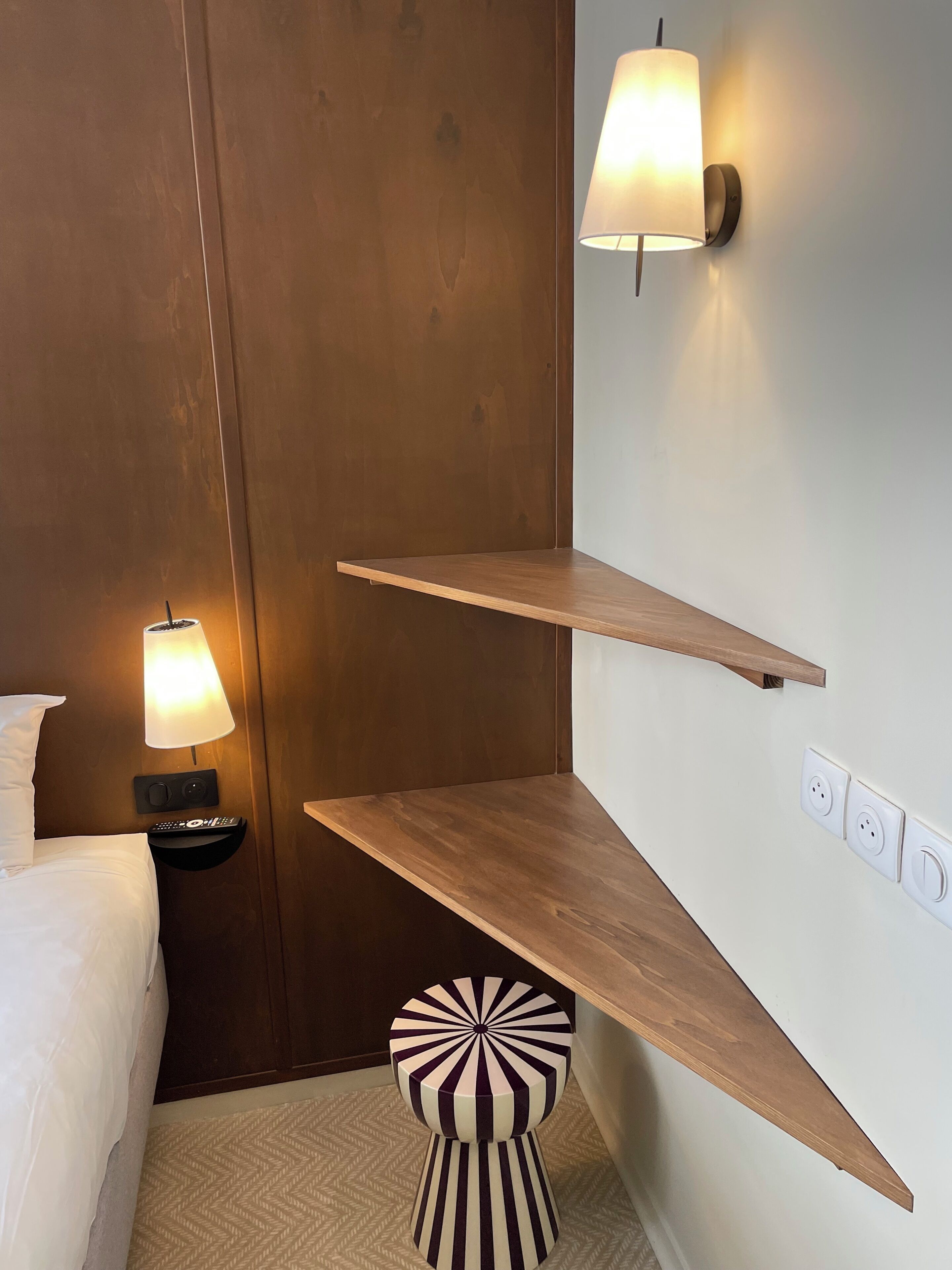 A hotel room with a bed, bedside lamp, wall-mounted shelf, and a unique striped ottoman.