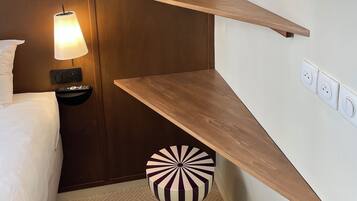 A hotel room with a bed, bedside lamp, wall-mounted shelf, and a unique striped ottoman.
