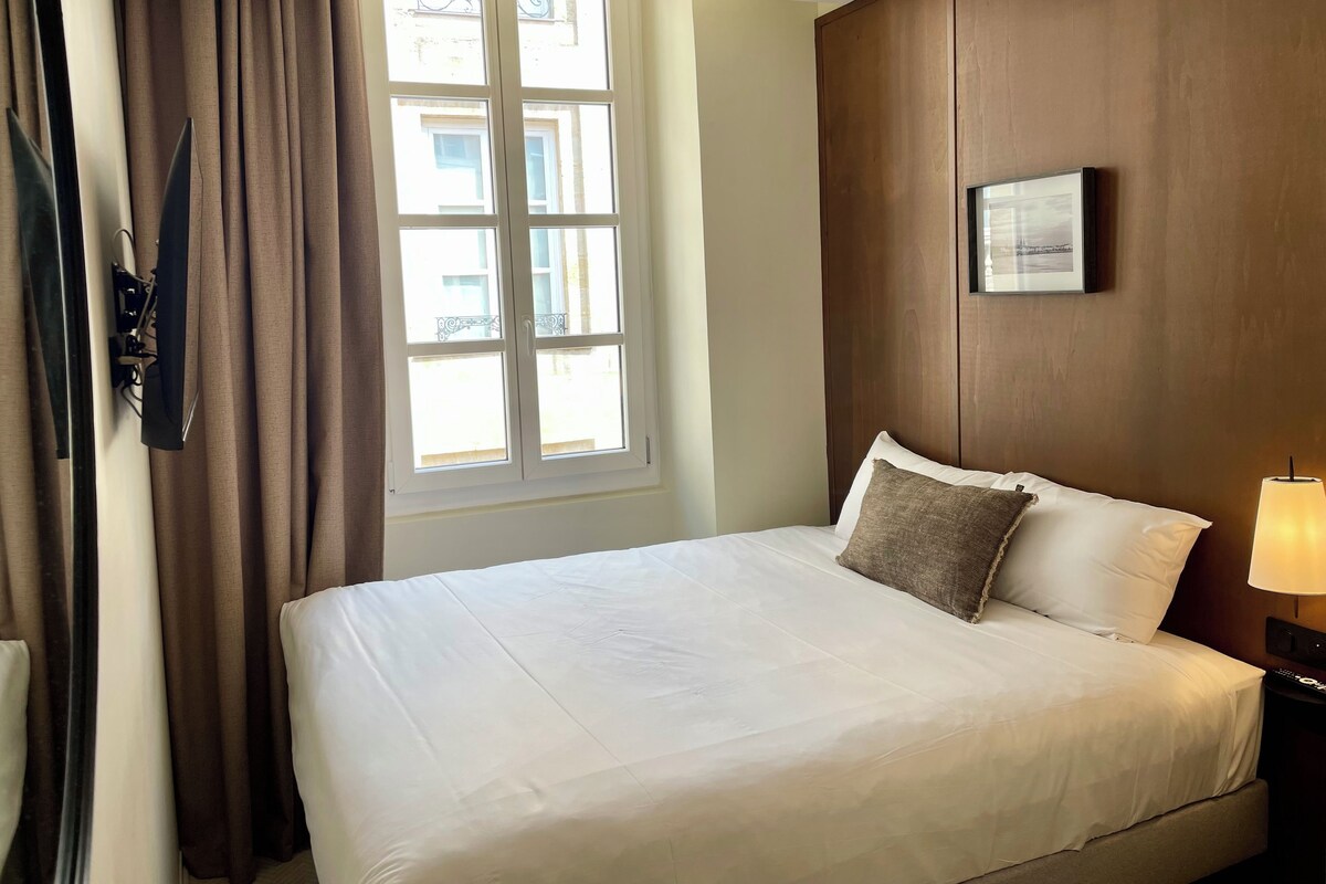 Una camera d'albergo con un letto, una finestra con tende, un quadro appeso al muro e una lampada da comodino.