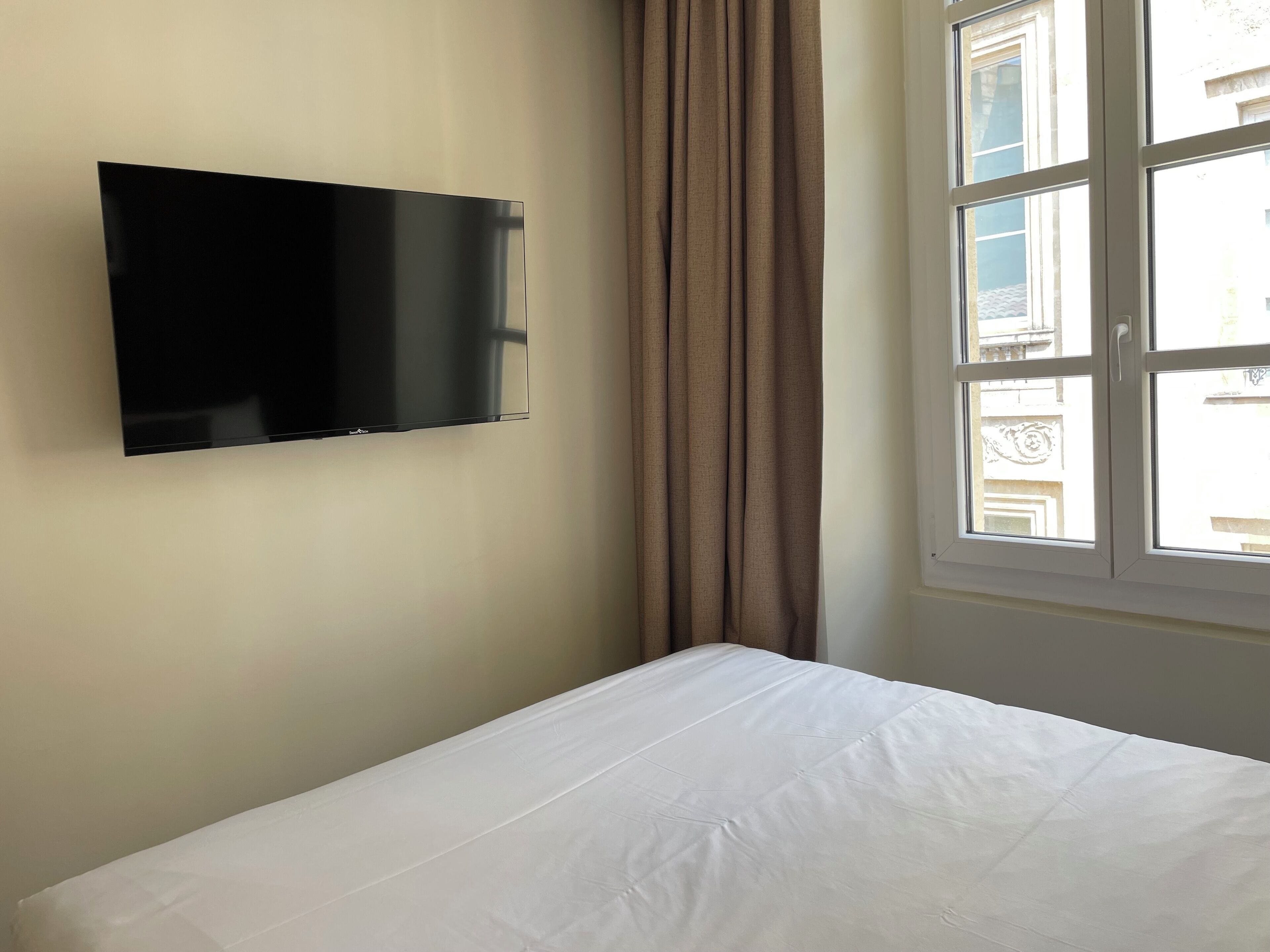 A room with a bed, a flat-screen TV mounted on the wall, and a window with curtains.