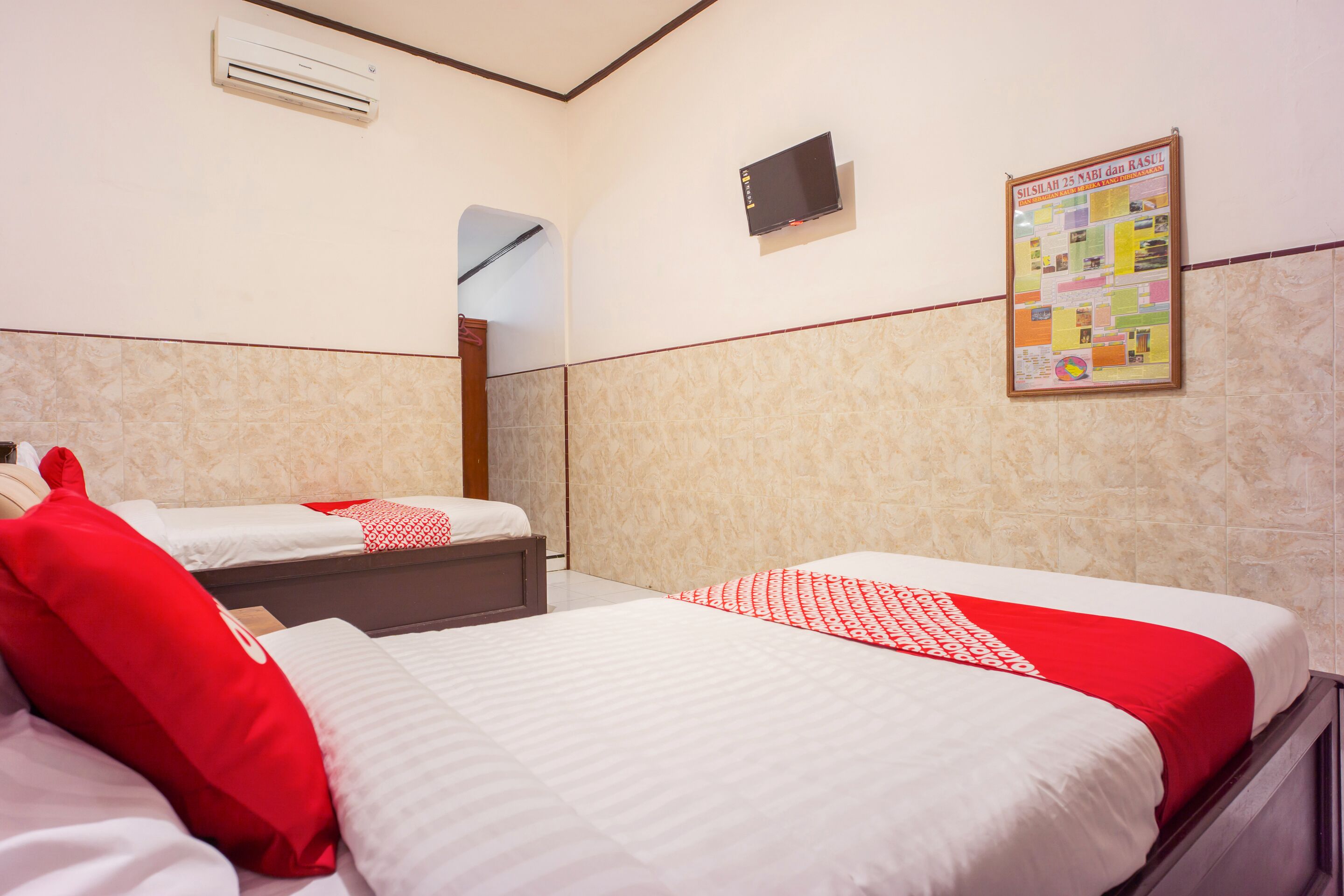 A hotel room with two beds, each with red and white bedding, a mounted TV, and a bulletin board on the wall.