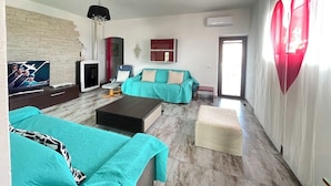 A modern living room with teal sofas, a coffee table, and a TV. There is a fireplace, a red wall decoration, and a large window with curtains. - H 51 FAMILY HOUSE  (Budoni)