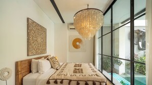 A modern bedroom with a large bed, a chandelier, and a view of an outdoor pool. - Maison Jolie Villa by Betterplace (Canggu)