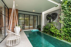 Un área de piscina moderna con una silla colgante, un mural de un elefante y abundante vegetación.