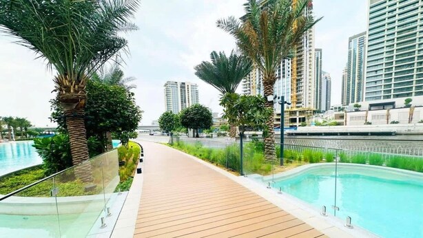 A wooden deck with a glass railing overlooking a swimming pool, surrounded by palm trees and modern buildings. - Silkhaus Surf 1 Dubai Creek Harbour (Dubai)
