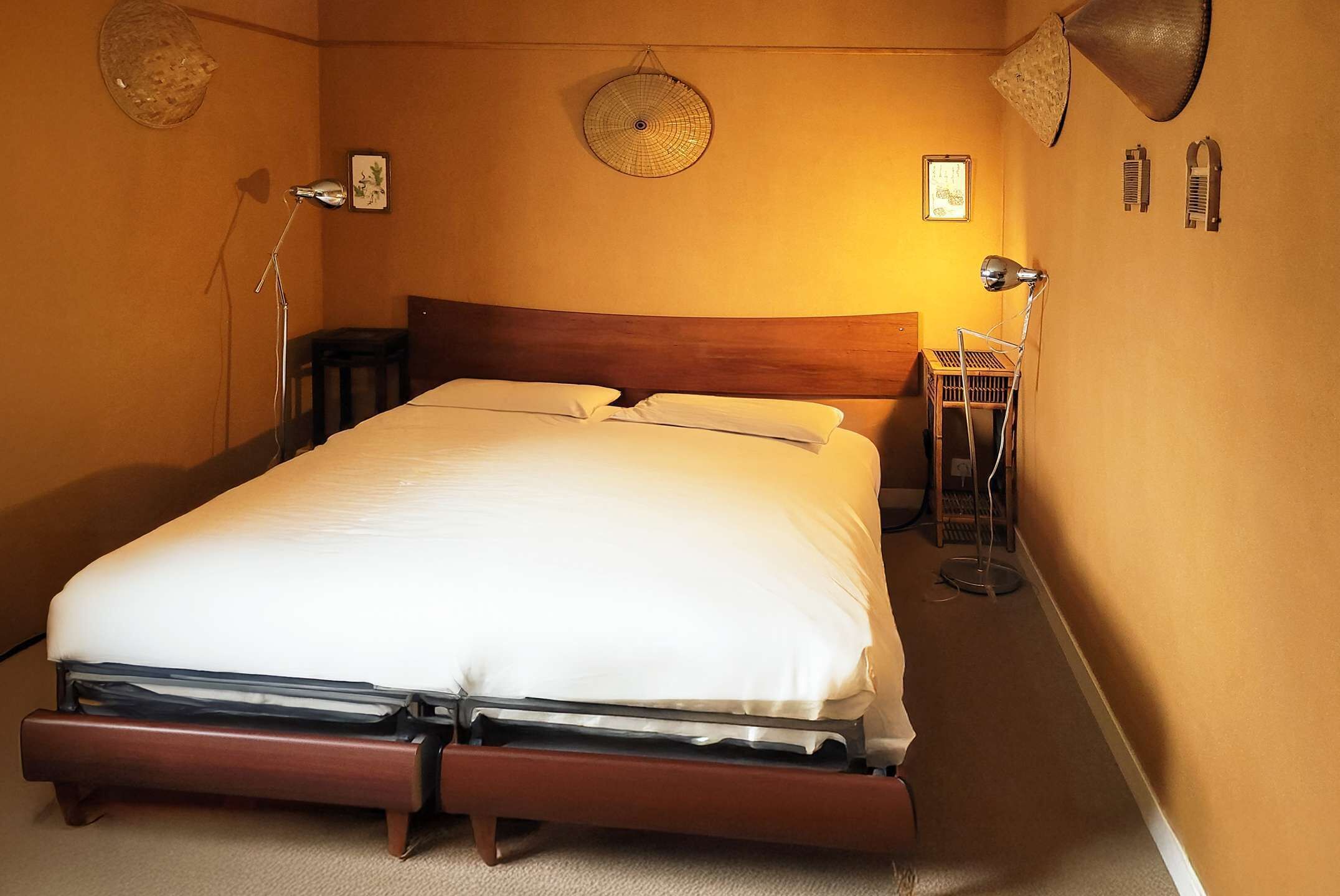 Una cama bien tendida en una habitación con cabecera de madera, una lámpara de pie y un ventilador de pared.