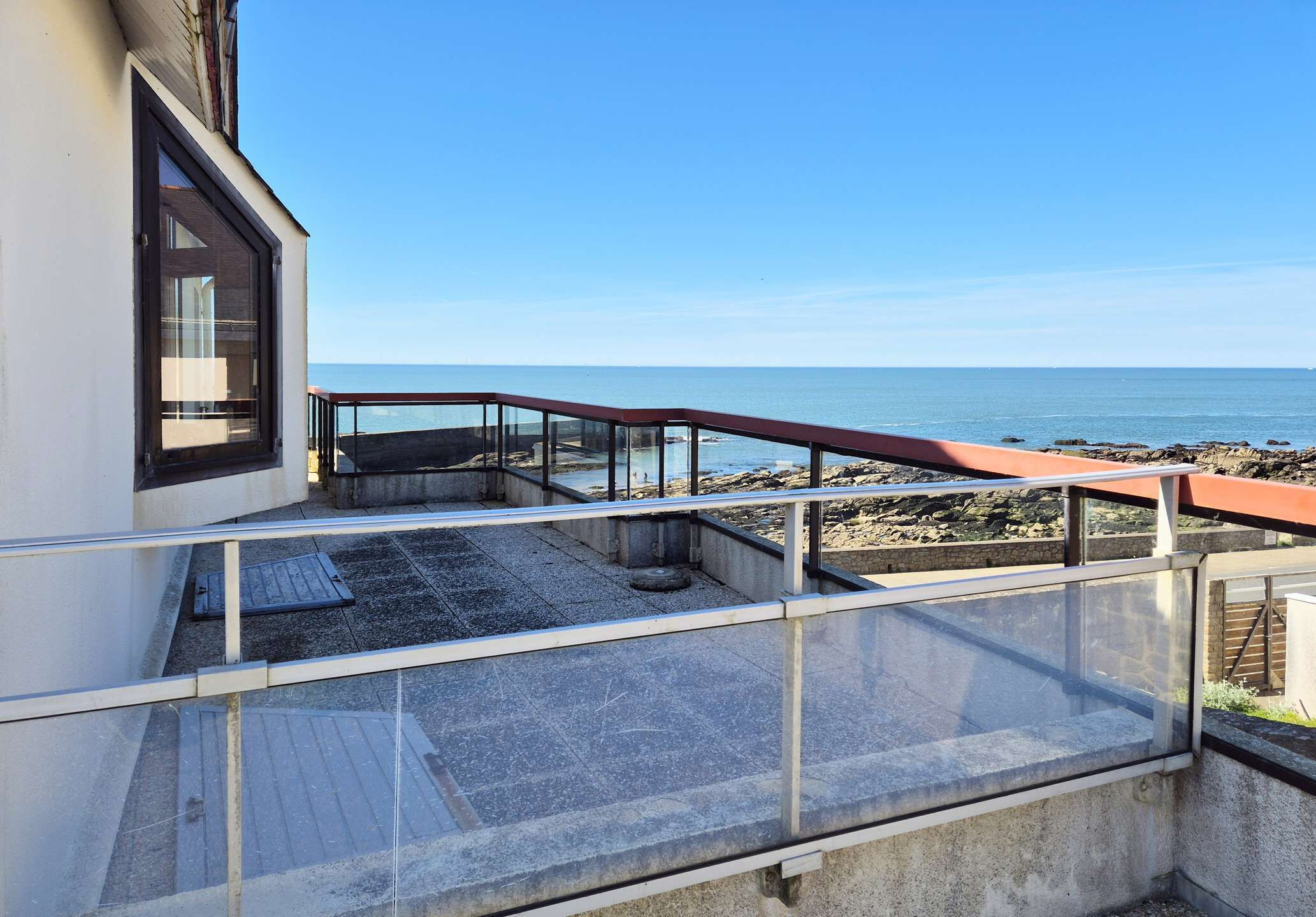 Un balcón con vista al mar, barandilla de cristal y piso de concreto.