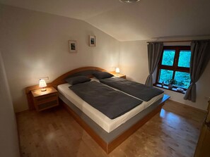 A bedroom with a large bed, two bedside tables with lamps, a window with curtains, and a wooden floor. - Ferienwohnung Haus am Wald (Marktschellenberg)