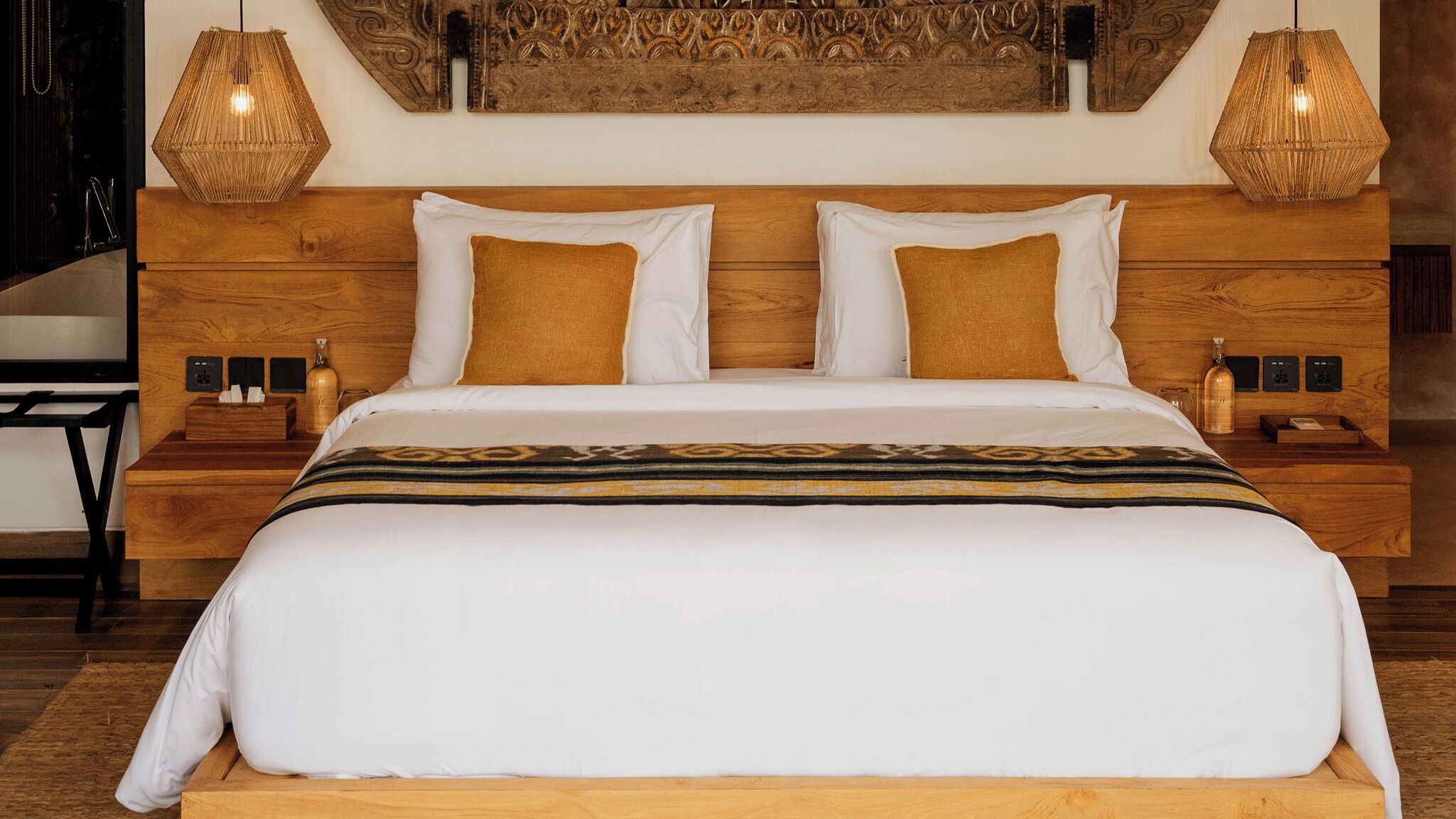 A neatly made bed with a white duvet, two brown pillows, and a patterned throw. There are two bedside tables with lamps, a wooden headboard, and a decorative wall piece above the bed.