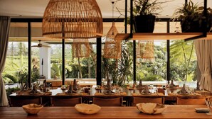 Comedor con mesas dispuestas para una comida, ventanas amplias con vistas a un jardín exuberante y lámparas colgantes tejidas.