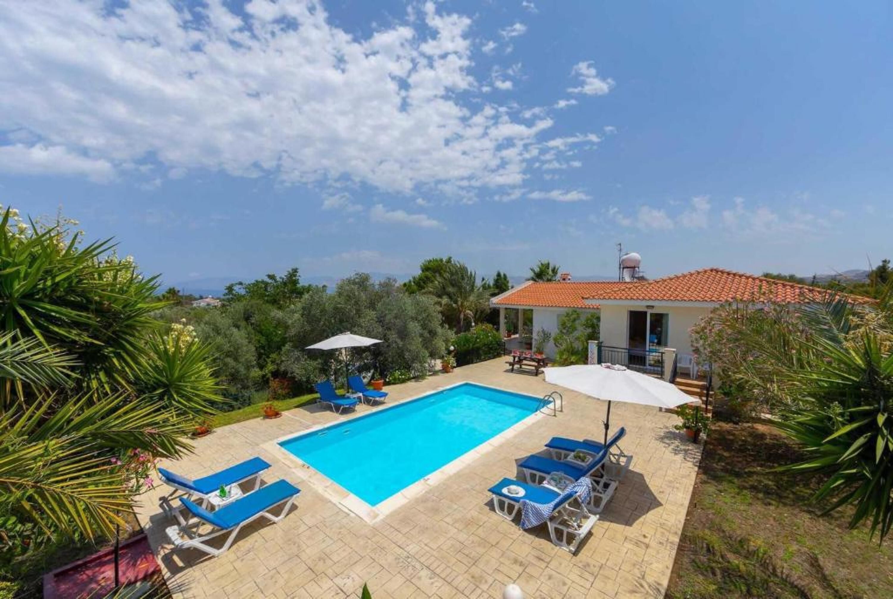 A swimming pool with lounge chairs and umbrellas, surrounded by greenery and a building with a red-tiled roof.