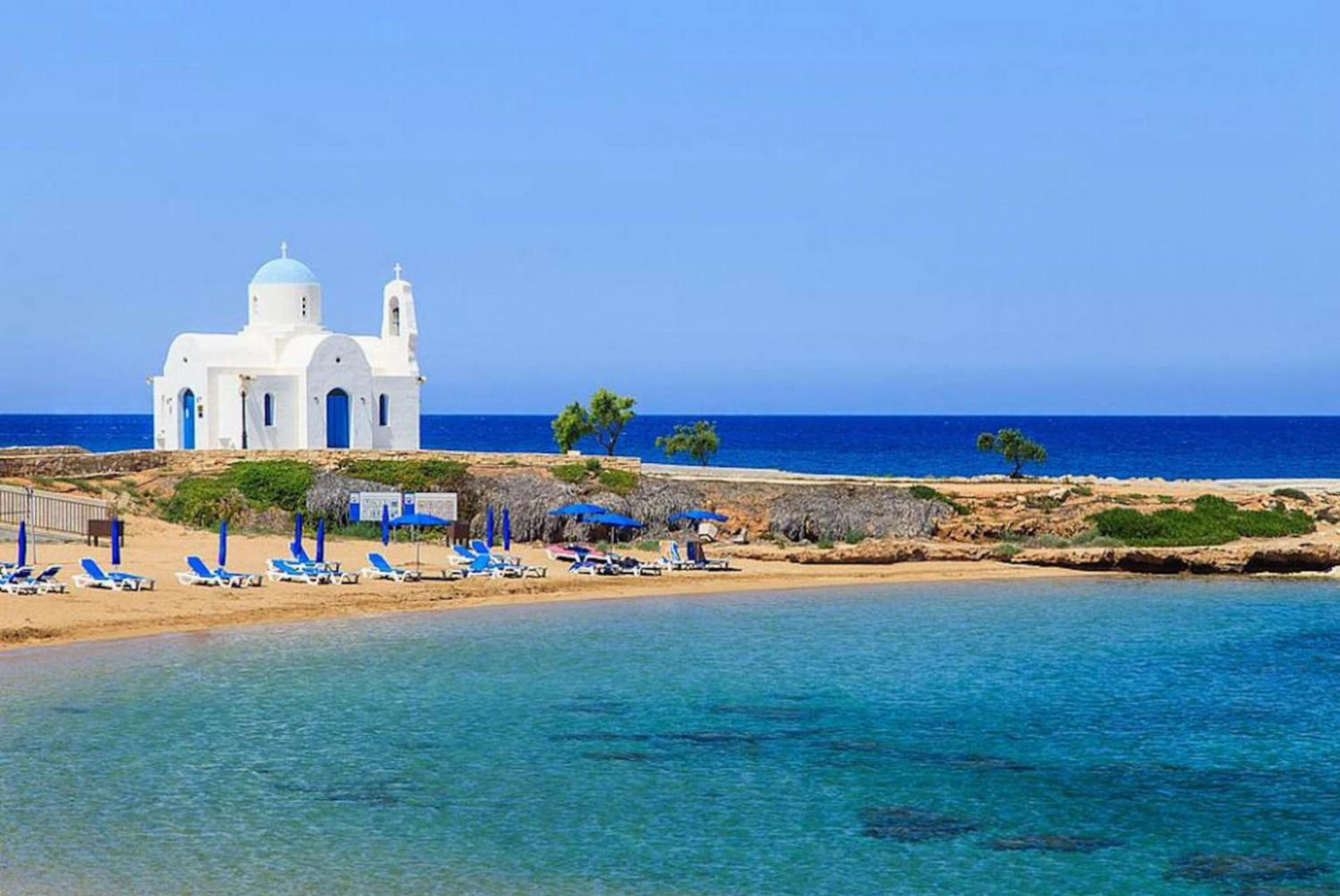 Una iglesia blanca con dos cúpulas y un campanario, una playa de arena con sombrillas azules, y el mar al fondo.