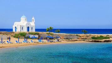 Una iglesia blanca con dos cúpulas y un campanario, una playa de arena con sombrillas azules, y el mar al fondo.
