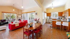A spacious living area with a red sofa, wooden dining table, and a fireplace. The kitchen includes wooden cabinets and a small island with bar stools. - Villa Katerina in Neo Chorio (Neo Chorio)