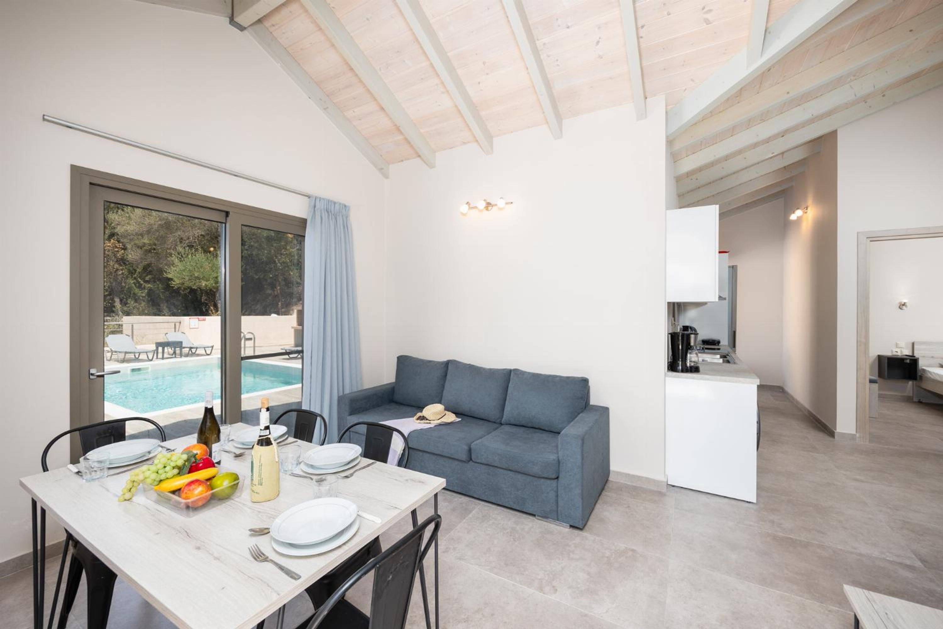 Une pièce moderne avec une table à manger dressée pour deux personnes, un canapé gris, un coin cuisine et une piscine visible par la fenêtre.