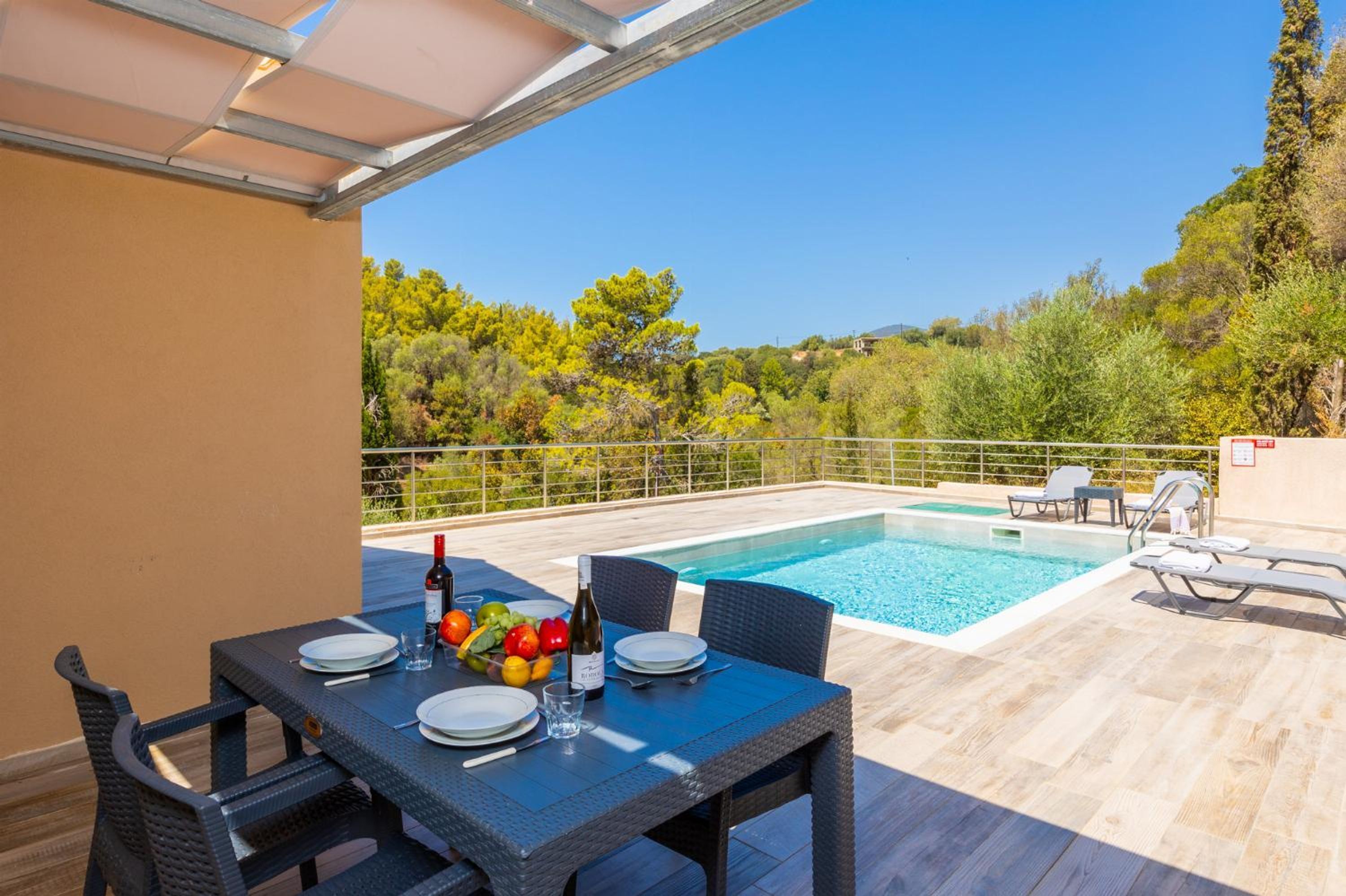 A poolside dining area with a table set for two, a bottle of wine, and a fruit bowl.