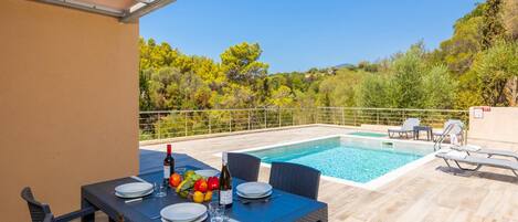 A poolside dining area with a table set for two, a bottle of wine, and a fruit bowl.