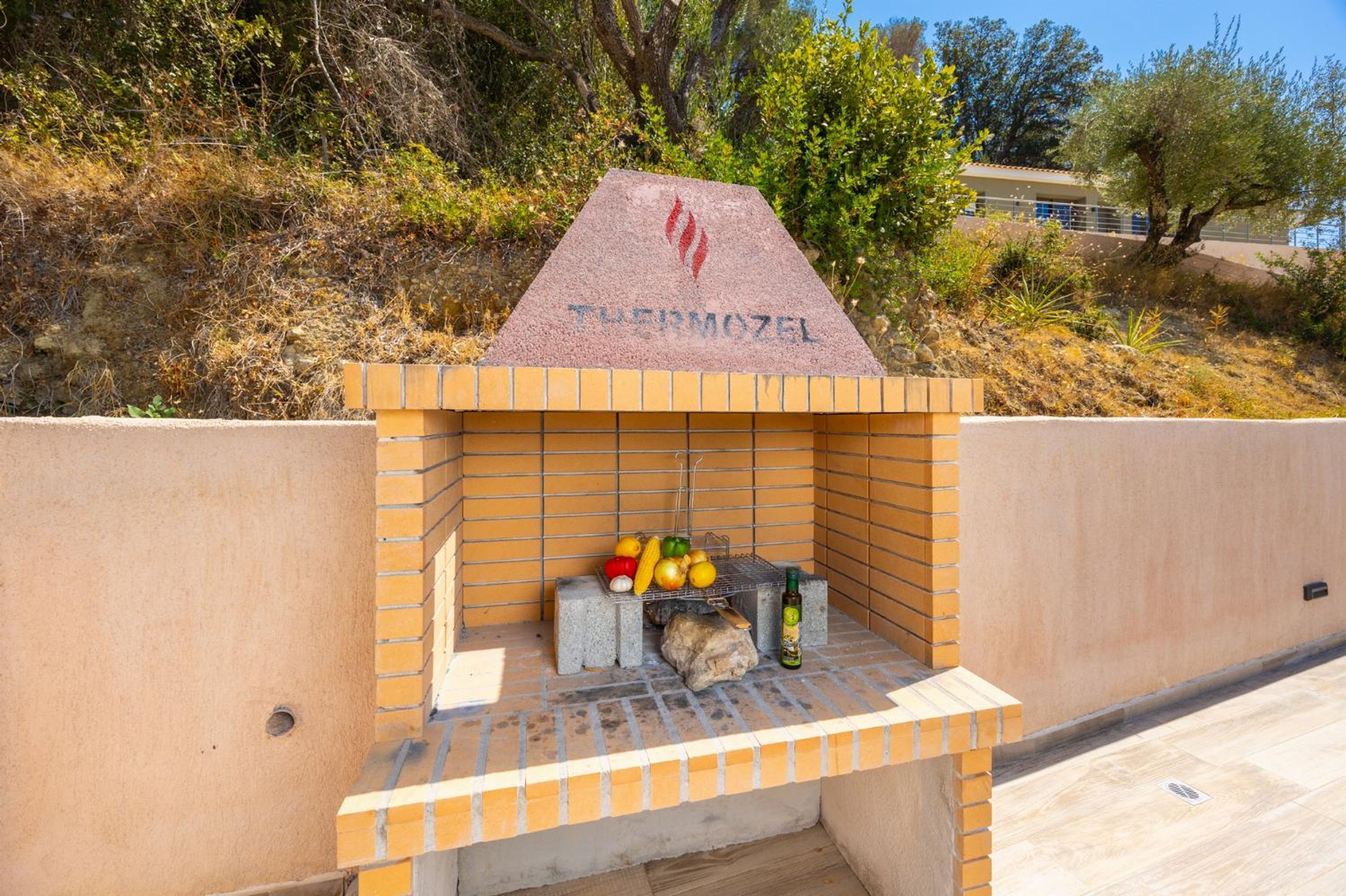 A brick barbecue grill with a stone sign above it, surrounded by a beige wall and some vegetation.