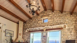 Una habitación rústica de paredes de piedra y techo de madera, con una mesa de comedor y sillas, una mesita redonda con un libro, un sofá y un televisor sobre un mueble de madera.