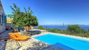 Piscina con suelo de piedra y sillones reclinables naranjas, rodeada de vegetación y vistas al mar.