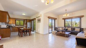 A spacious living area with a dining table and chairs, a sofa, and a coffee table. There is a kitchen with wooden cabinets and a refrigerator. Large windows provide natural light and views of the outdoors. - Villa Kambos Dio in Polis (Chrysochou)