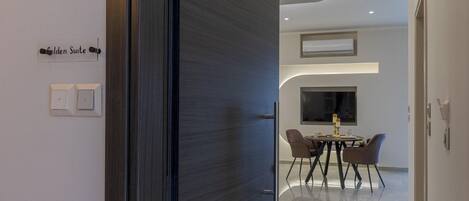 Moderni hotellihuone, jossa on tumma puuovi, kyltti, jossa lukee ”Golden Suite”, ruokailutila pyöreällä pöydällä ja tuoleilla sekä suuri ikkuna.