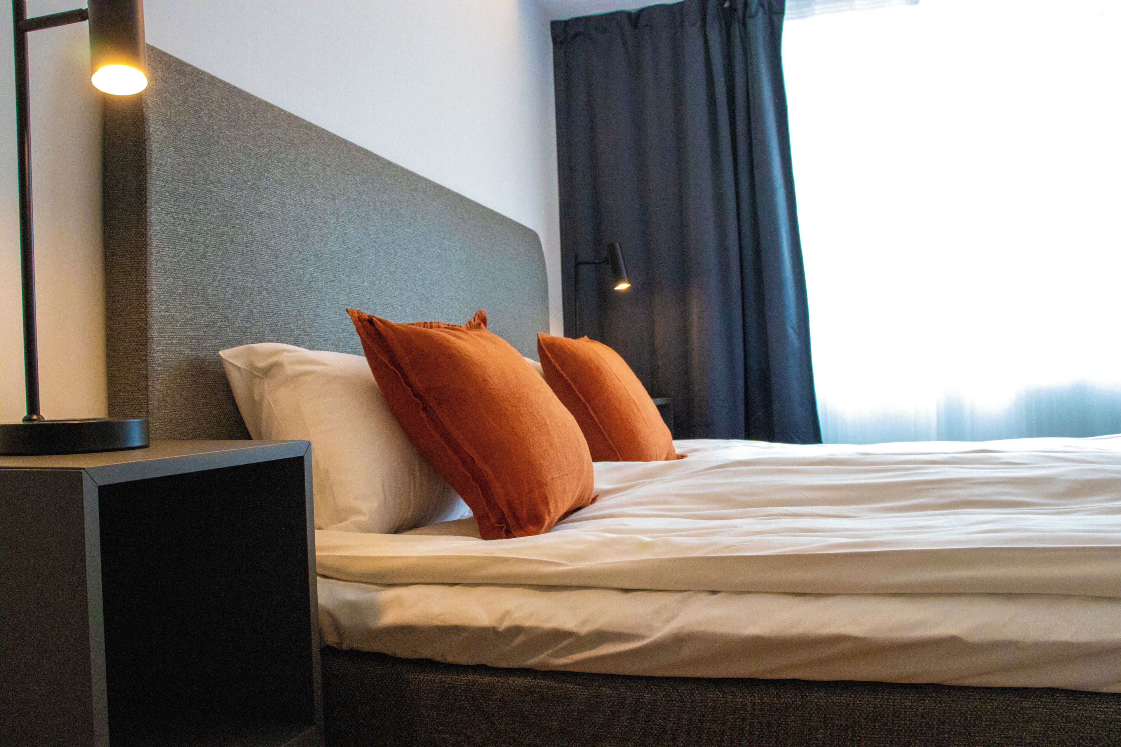 Una cama bien hecha con sábanas blancas y dos almohadas naranjas, una mesita de noche con lámpara y una cortina oscura parcialmente abierta.