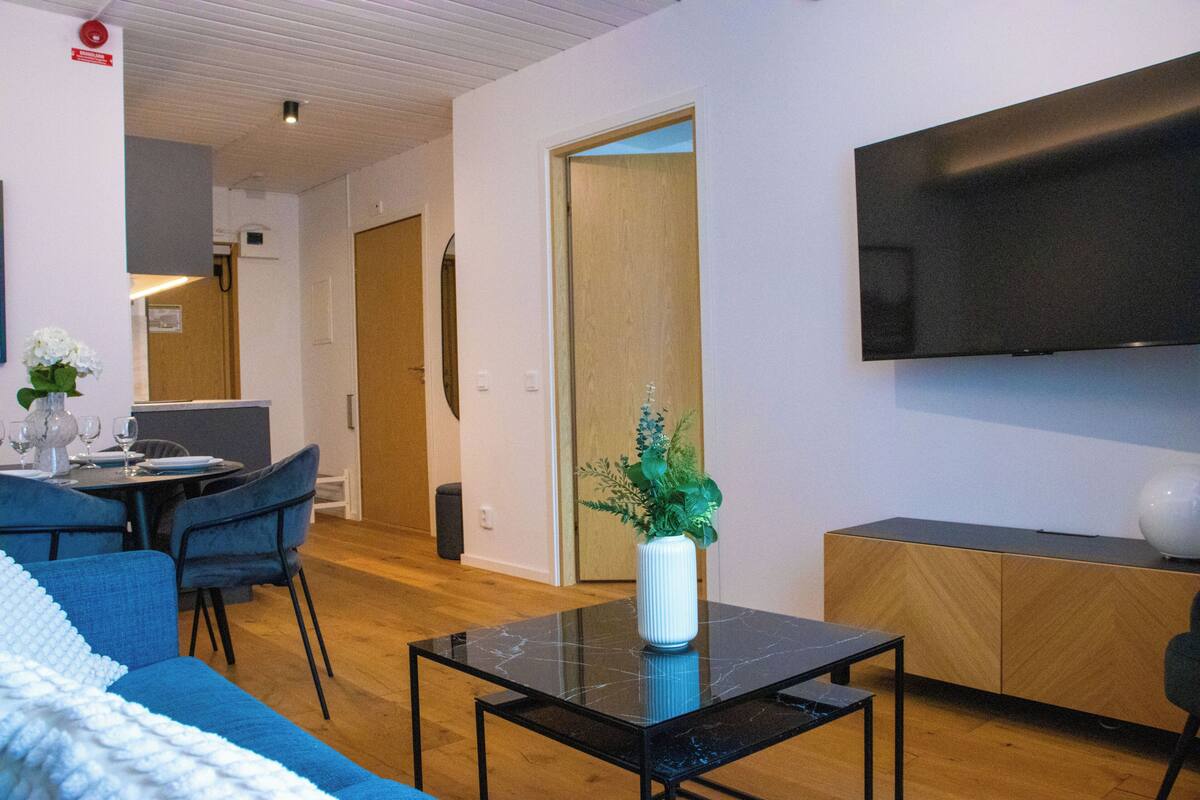 Una habitación de hotel moderna con comedor, televisión de pantalla plana, aparador de madera y un florero con planta.