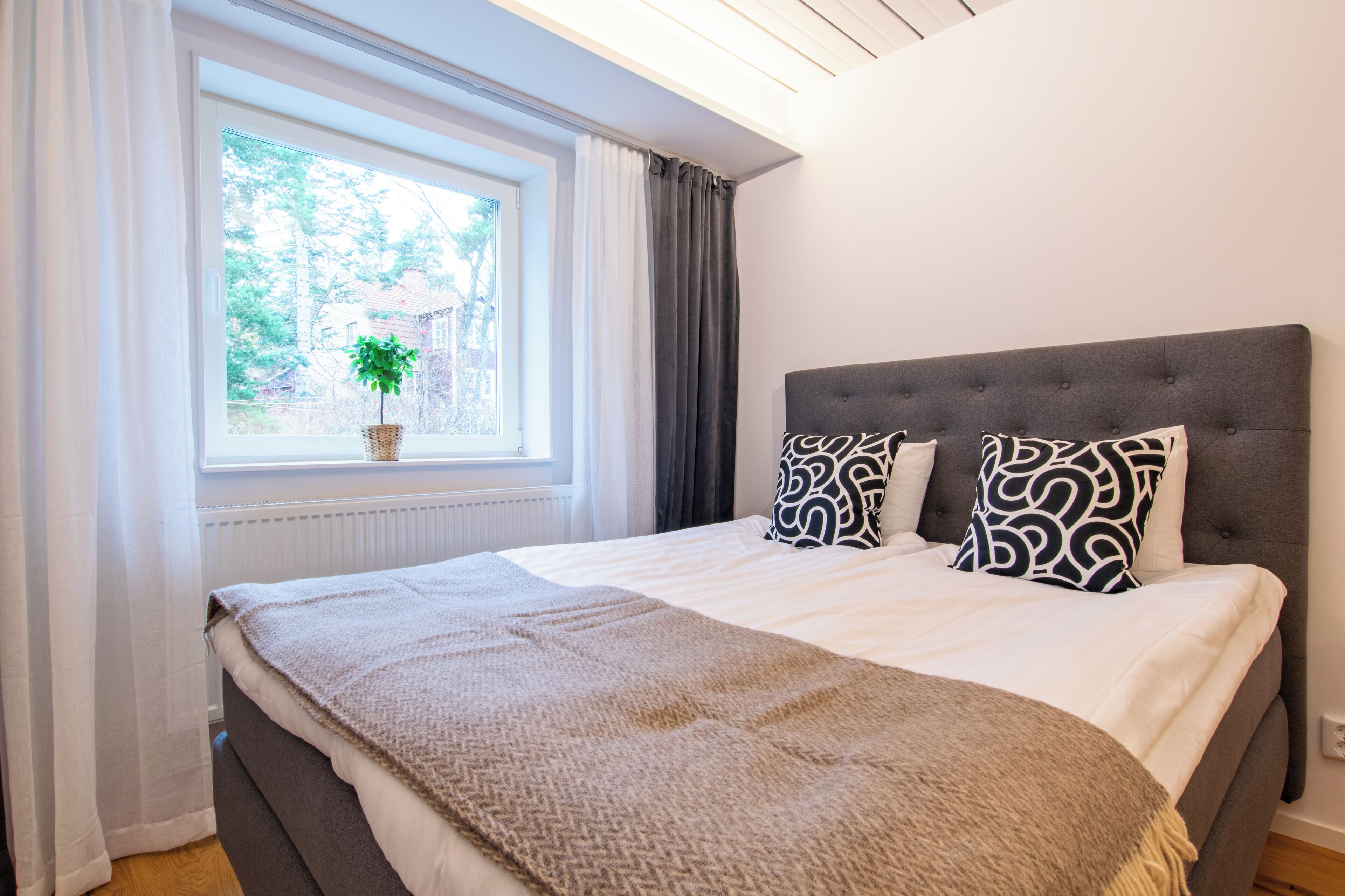 Un dormitorio con una cama grande, una ventana con cortinas, una planta en maceta y un cabecero tapizado en gris.