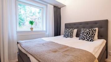 Un dormitorio con una cama grande, una ventana con cortinas, una planta en maceta y un cabecero tapizado en gris.