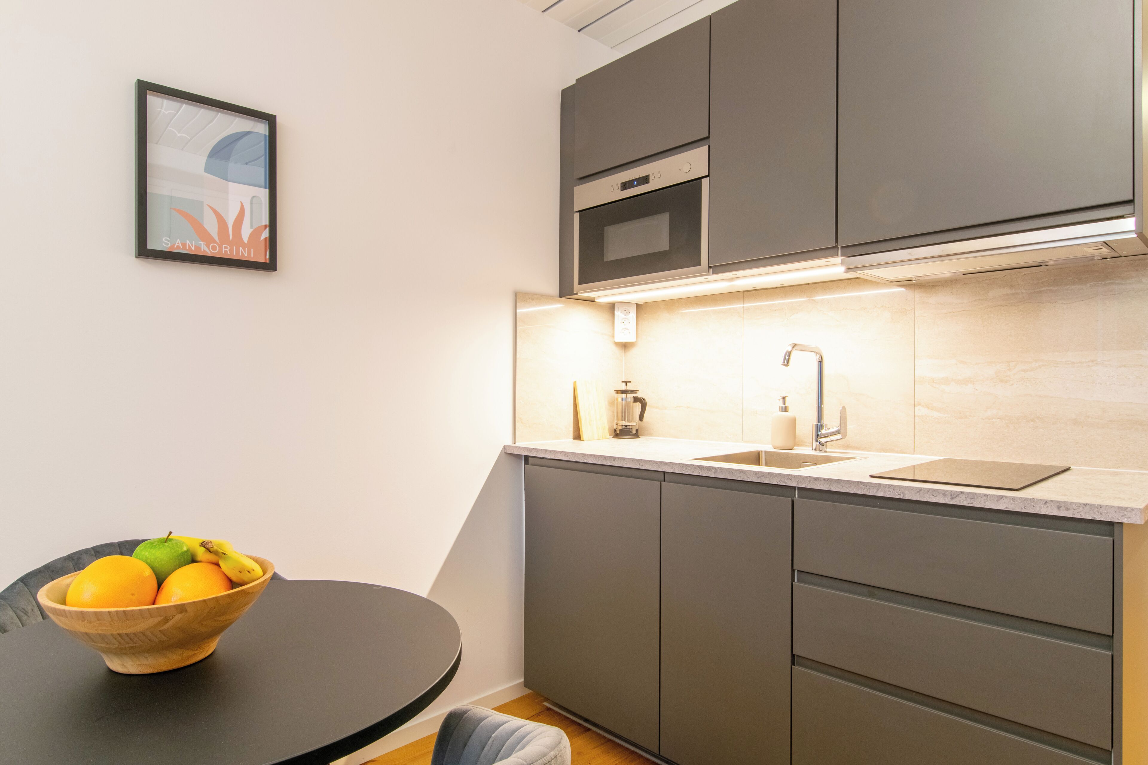 Een moderne keuken met grijze kastjes, een ingebouwde oven en een spoelbak. Op een ronde tafel staat een schaal met fruit.