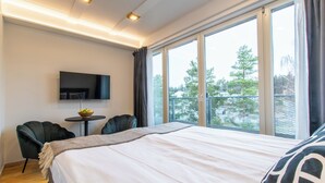 Una habitación de hotel moderna con una cama grande, un televisor de pantalla plana, una pequeña mesa con un cuenco de fruta, dos sillas y un balcón con vistas a árboles y edificios.