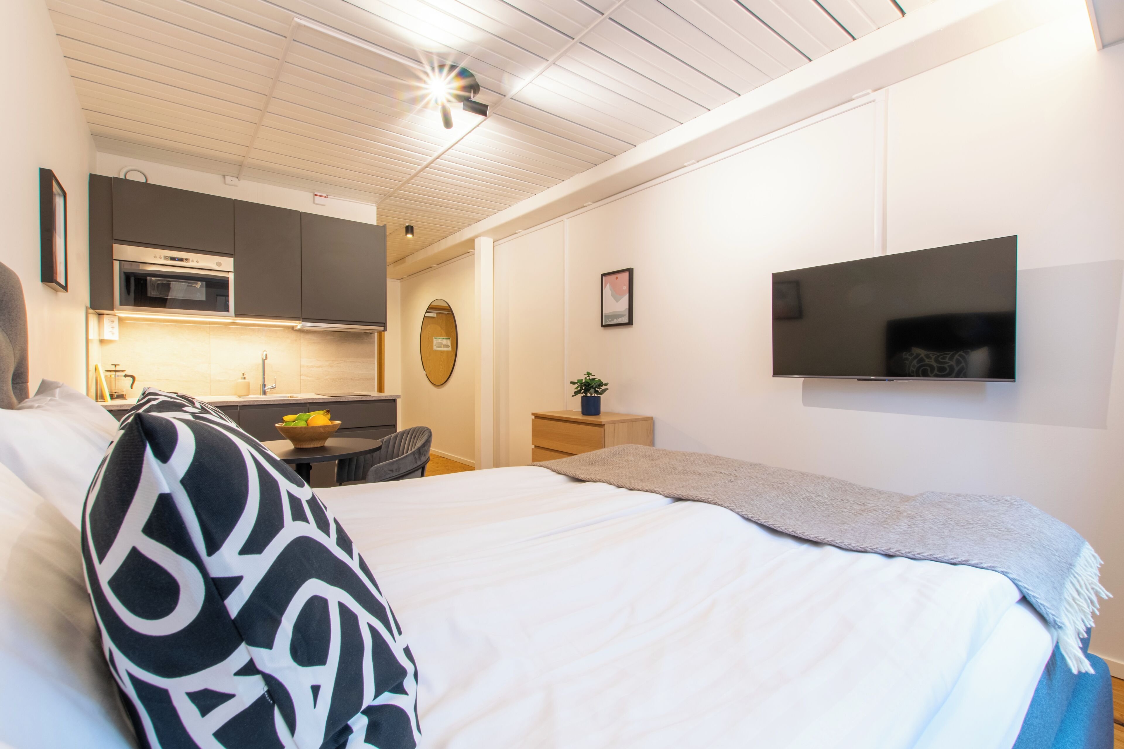 A hotel room with a bed, a TV mounted on the wall, a kitchenette, and a small desk with a chair.