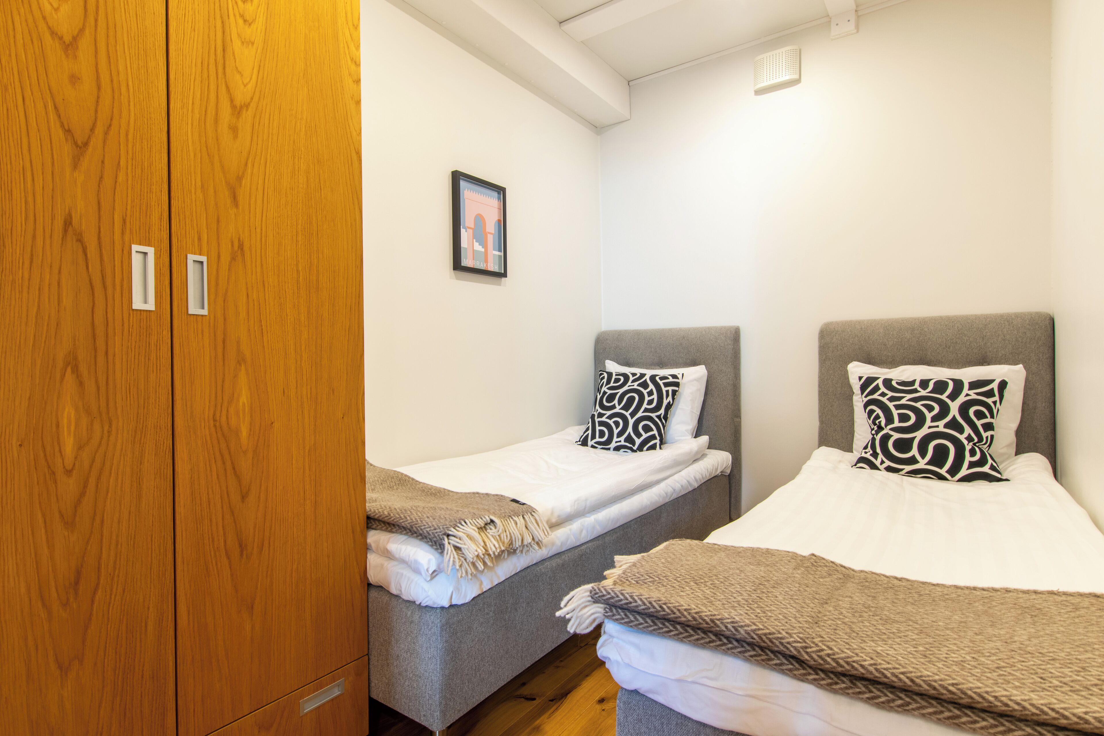 A room with two single beds, each with patterned pillows and a blanket. There is a wooden wardrobe and a framed picture on the wall.