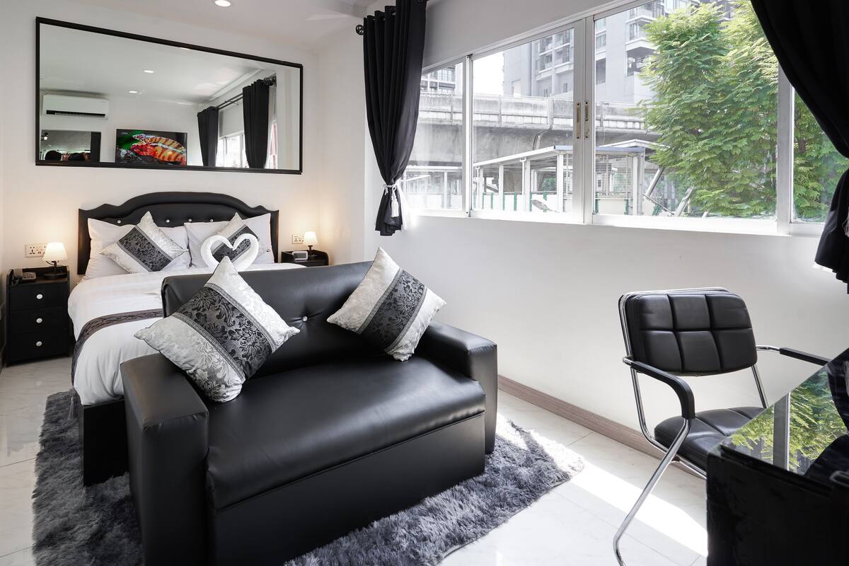 Quarto moderno com sofá de couro preto, cama com almofadas estampadas, mesa lateral com luminária e uma grande janela com vista para prédios e árvores.